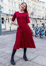 Stylish Mum with dark hair pinned up. She has her hand on her hip and has a huge smile. She is wearing a stunning red midi dress which has zips for breastfeeding. She is wearing black tights and boots. The dress is made by Stylish Mum who makes breastfeeding dresses in the UK. 