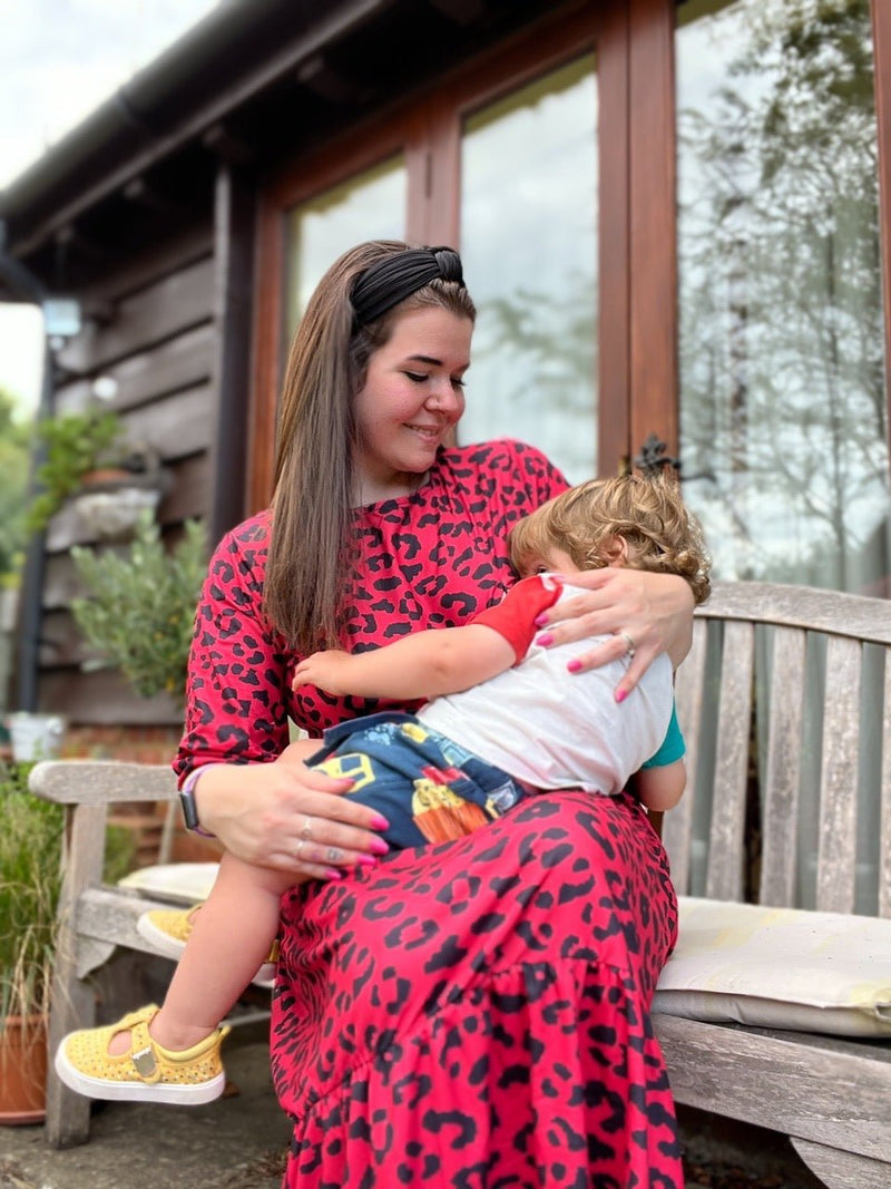 Mum with long dark hair sat on bench outside breastfeeding her toddler. She is wearing a beautiful red leopard print long midi dress. The dress has hidden zips for breastfeeding so although the mum is breastfeeding it is very discreet. The dress is a best seller by Stylish Mum which is a breastfeeding UK brand. 